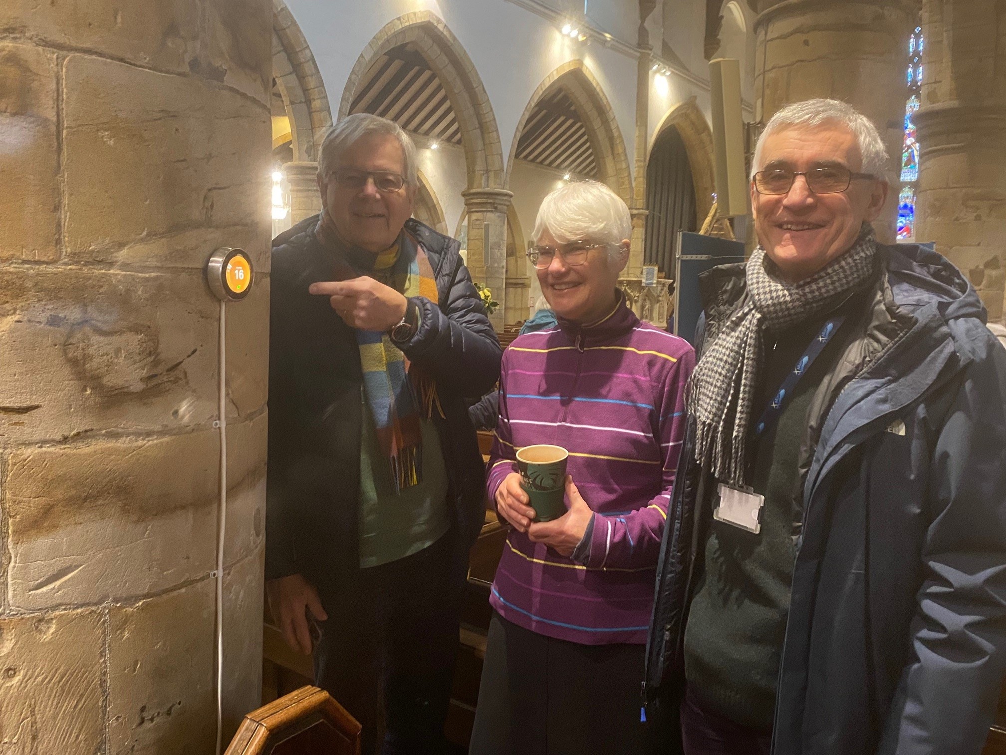 Picture of 3 of the members of the church smiling at the nest thermostat installed on the church wall
